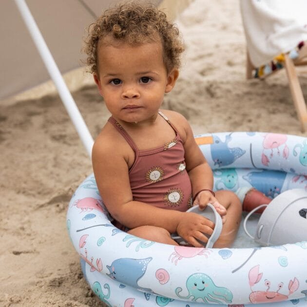 Maluch z kręconymi włosami w brązowym kostiumie kąpielowym siedzi w dmuchanym pierścieniu Basenik Sea Animals 60 cm na piaszczystej plaży - idealny na akcesoria do basenów dla dzieci w naszym internetowym butiku ogrodniczym.