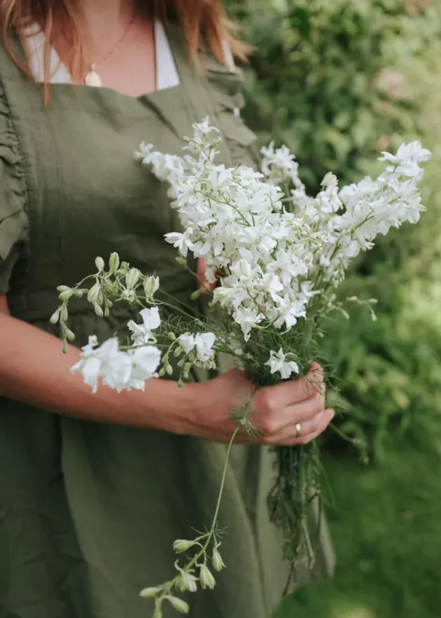 Kolekcja Gardening Boutique: Osoba w zielonym fartuchu trzymająca wykwintny biały bukiet kwiatów na tle bujnego ogrodu.