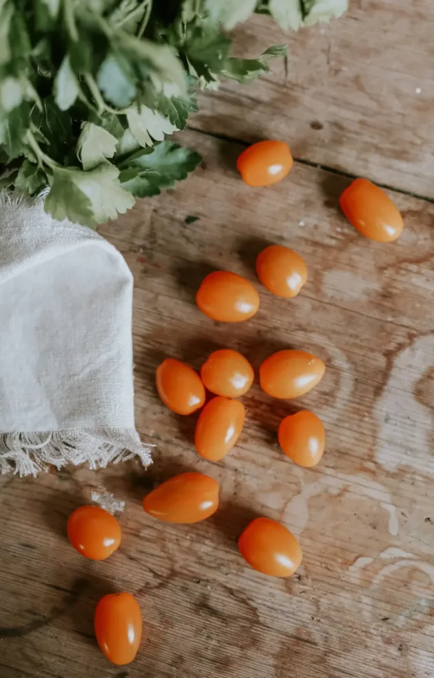 Świeże, intensywnie pomarańczowe pomidorki koktajlowe rozrzucone na drewnianym stole obok bujnej wiązki zielonych liści, artystycznie przykryte elegancką białą tkaniną - idealne do Twojego ekologicznego ogrodu warzywnego w naszym internetowym butiku ogrodniczym.