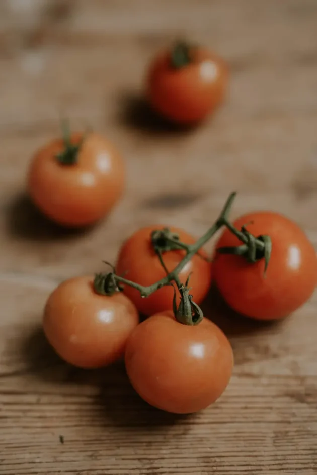 Grono dojrzałych, czerwonych pomidorów Sungold na krzaku w internetowym butiku ogrodniczym, spoczywające na rustykalnej drewnianej powierzchni z rozmytymi pomidorami w tle, tworzące atmosferę ogrodu, niczym ze świeżego gospodarstwa.