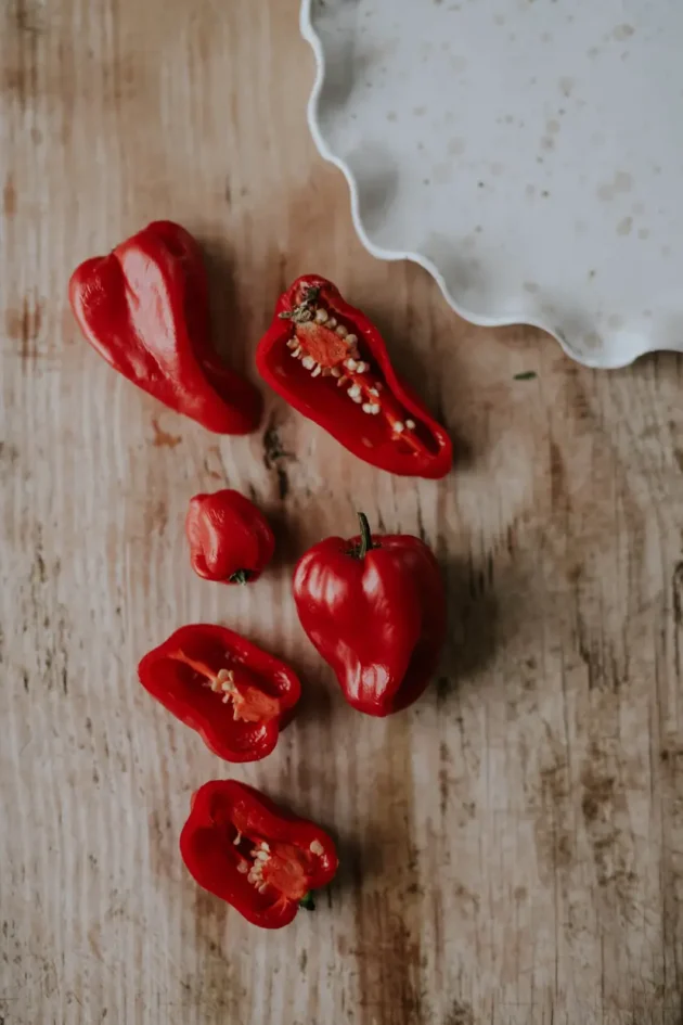 Żywe, różnorodne czerwone papryczki chili Habanero Red na rustykalnym drewnianym tle - internetowy butik ogrodniczy. Całe i połówki papryczek chili z nasionami na falistym białym talerzu w rogu. Idealne dla miłośników pikantnych ogrodów!.