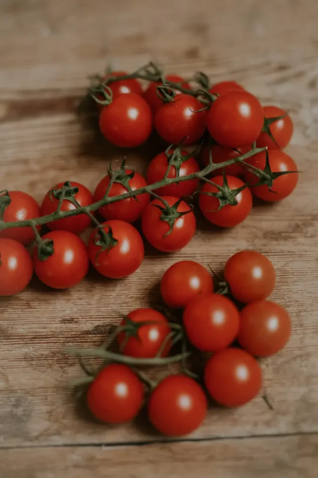 Żywy kiść świeżych czerwonych pomidorków koktajlowych Sweetie na winorośli wystawionych na rustykalnych drewnianych deskach. Idealny dla kupujących w internetowych butikach ogrodniczych, którzy szukają produktów ekologicznych klasy premium. Małe, soczyste i okrągłe pomidory z kilkoma rozrzuconymi luźnymi pomidorami dodają autentycznego charakteru z ogrodu na stół. Idealny do wzbogacenia domowej kolekcji ogrodowej lub przepisów kuchennych.