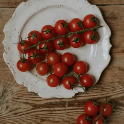 Ulepsz swój ogród przydomowy za pomocą żywych czerwonych pomidorków koktajlowych Sweetie z naszego internetowego butiku ogrodniczego. Te soczyste pomidory z bujnymi zielonymi łodygami, umieszczone na uroczym rustykalnym drewnianym stole, elegancko spoczywają na białym talerzu z falistymi krawędziami. Jeden dodatkowy bukiet umieszczony obok talerza podkreśla piękny kontrast z wyblakłą fakturą stołu. Idealne do dodawania koloru i świeżości do Twoich kulinarnych kreacji lub ogrodowych ekspozycji!.