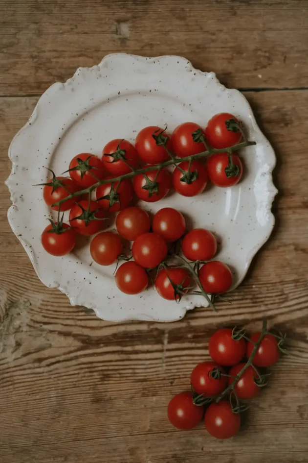 Ulepsz swój ogród przydomowy za pomocą żywych czerwonych pomidorków koktajlowych Sweetie z naszego internetowego butiku ogrodniczego. Te soczyste pomidory z bujnymi zielonymi łodygami, umieszczone na uroczym rustykalnym drewnianym stole, elegancko spoczywają na białym talerzu z falistymi krawędziami. Jeden dodatkowy bukiet umieszczony obok talerza podkreśla piękny kontrast z wyblakłą fakturą stołu. Idealne do dodawania koloru i świeżości do Twoich kulinarnych kreacji lub ogrodowych ekspozycji!.