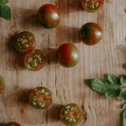 Świeże pomidorki koktajlowe Black Cherry, całe i przekrojone na pół, wystawione na rustykalnej drewnianej powierzchni z żywymi zielonymi liśćmi w czarującym ogrodowym butiku. Idealne dla Twojej ekologicznej inspiracji ogrodniczej.