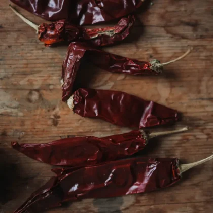 Pięć organicznych suszonych czerwonych papryczek chili Naga Black z łodygami, umieszczonych na rustykalnej drewnianej powierzchni – idealne rozwiązanie dla smakoszy i miłośników przypraw w naszym internetowym butiku ogrodniczym.