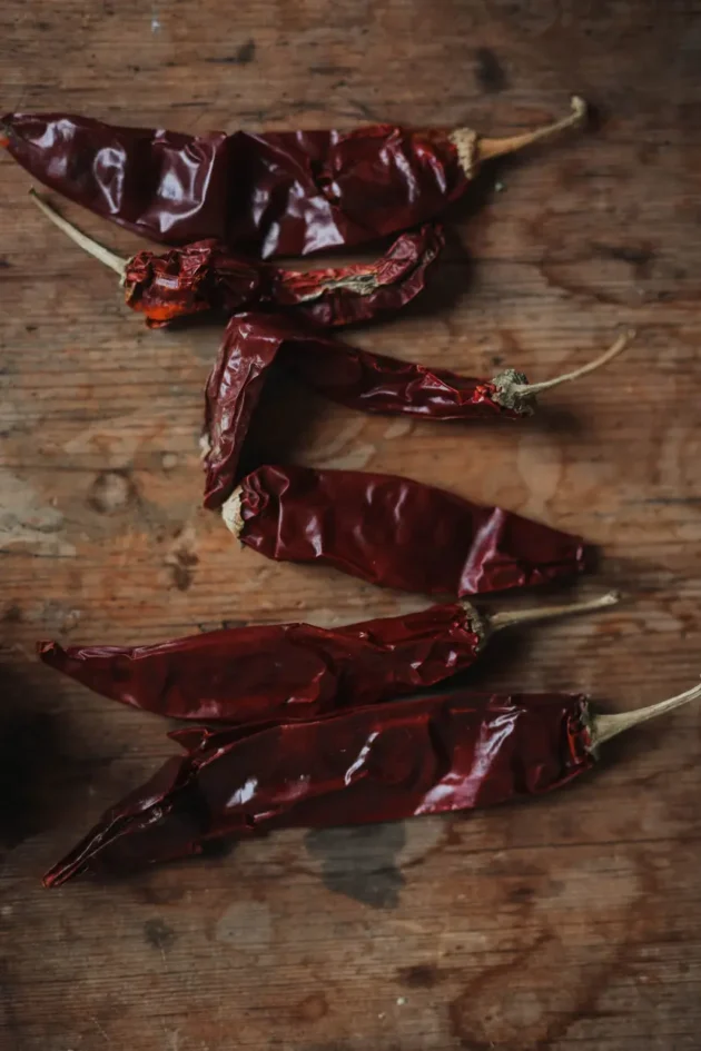 Pięć organicznych suszonych czerwonych papryczek chili Naga Black z łodygami, umieszczonych na rustykalnej drewnianej powierzchni – idealne rozwiązanie dla smakoszy i miłośników przypraw w naszym internetowym butiku ogrodniczym.