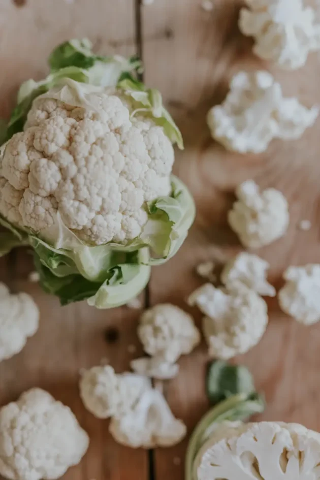 Zbliżenie świeżego, organicznego kalafiora Amazing z żywymi zielonymi liśćmi na rustykalnej drewnianej powierzchni, otoczonego rozproszonymi różyczkami kalafiora. Idealne dla internetowego butiku ogrodniczego prezentującego wysokiej jakości produkty z domowej uprawy i naturalny styl życia.