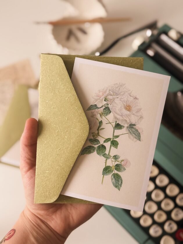 Ręka trzymająca botaniczną kartkę z pozdrowieniami „Biała Róża” z ilustracją kwiatową, częściowo wyjętą z zielonej koperty. W tle zabytkowa zielona maszyna do pisania i inspirowane ogrodem narzędzia do pisania — idealne dla miłośników roślin i pomysłów na prezenty ogrodnicze z internetowego butiku ogrodniczego.