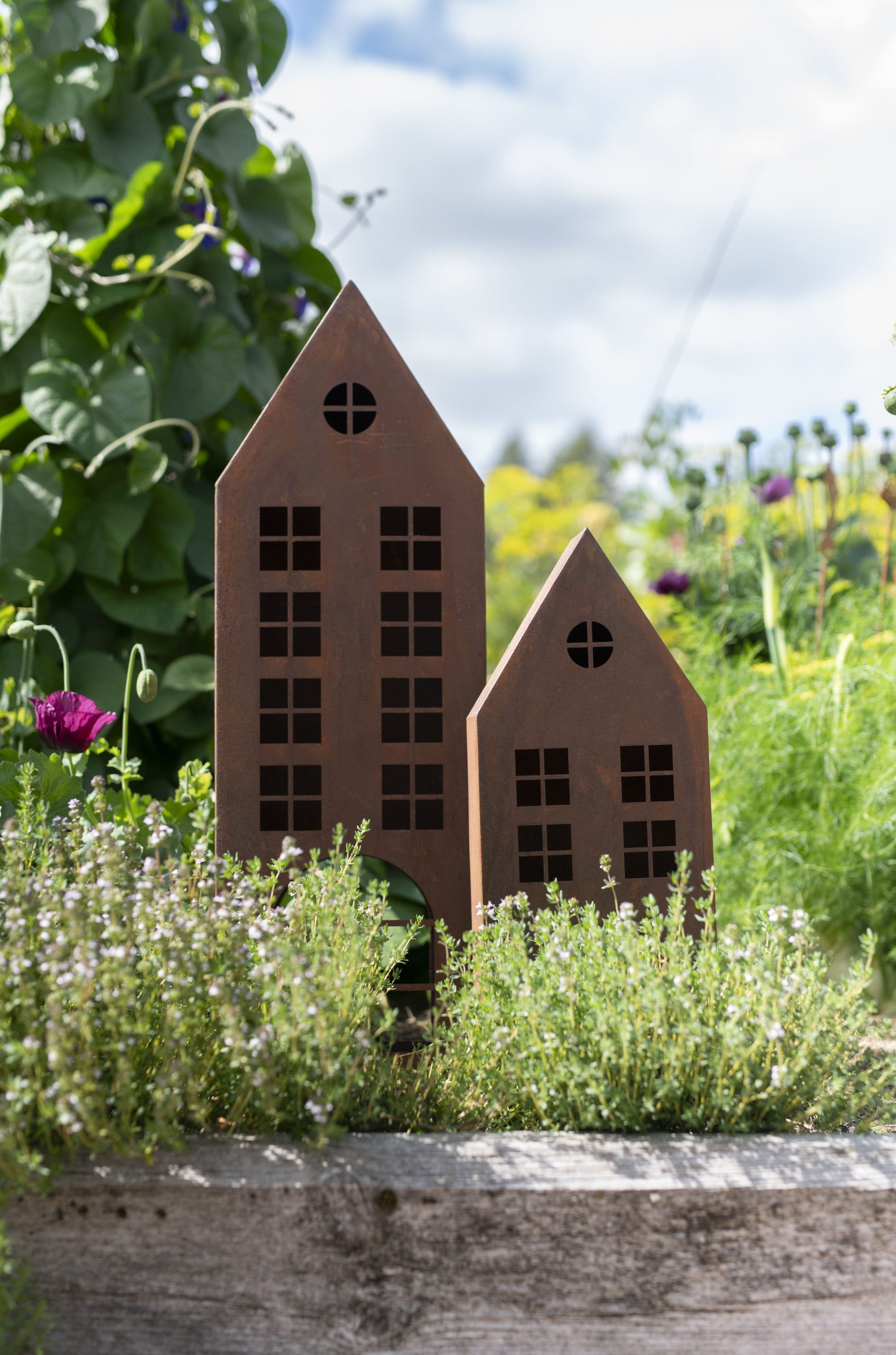 Dekoracyjne metalowe ozdoby z zestawu metalowych domków w bujnym ogrodzie kwiatowym, zieleni i kwiatach pod zachmurzonym niebem.