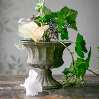 Metalowa doniczka Hilda S z zielonym bluszczem, kremowym kwiatem, nakrapianym piórkiem; rustykalne drewno i liściaste tło - dekoracja ogrodu.
