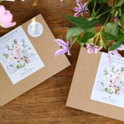 Kolekcja nasion 'New Cottage Garden' z kwiatowymi wzorami na drewnianym stole, otoczona różowymi i fioletowymi kwiatami ogrodowymi.