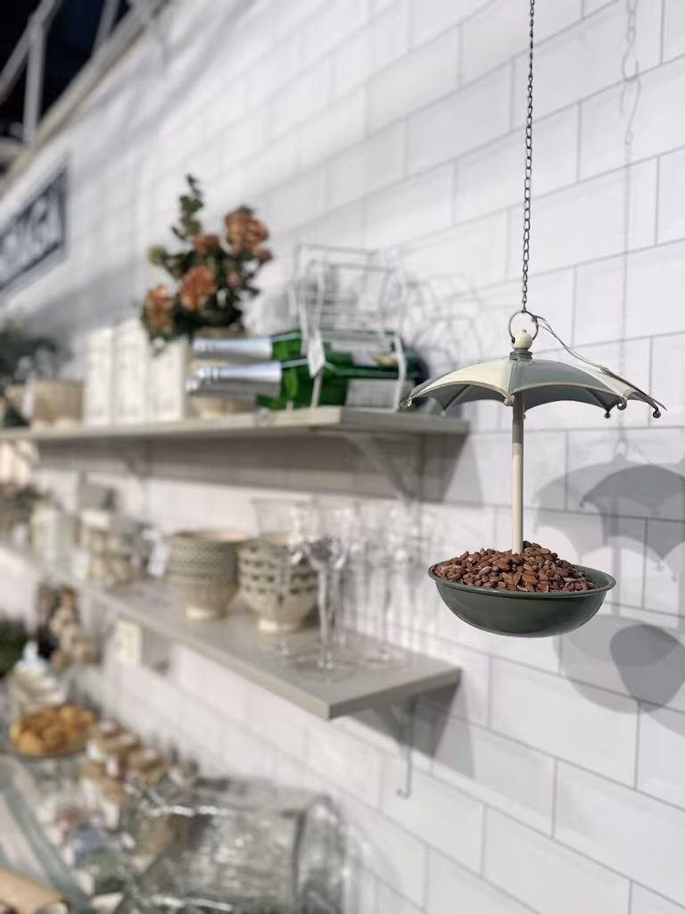 Karmnik/poidełko na ptaki Parasol - stylowa wisząca dekoracja ogrodowa - na tle niewyraźnych szklanych półek i białej ściany wyłożonej kafelkami.