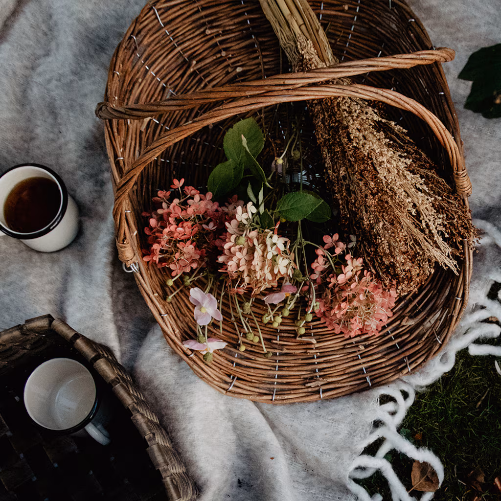 Kosz wiklinowy Iris z różowymi kwiatami i suszonymi roślinami na kocu, filiżanka do kawy, mały koszyk, trawa i biały szalik z dzianiny.