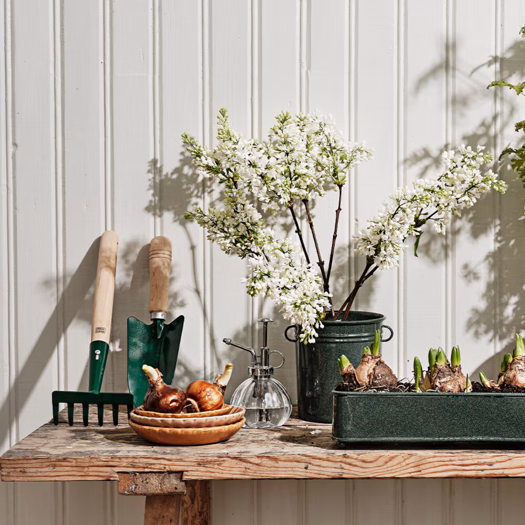 Drewniana dekoracja stołu z Drewnianym dziadkiem do orzechów, narzędziami ogrodniczymi, szklaną butelką do zamgławiania, cebulkami i białymi gałązkami kwiatowymi.