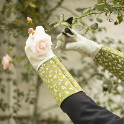 Ogrodnik w zielonych rękawiczkach w kwiaty wykorzystuje Małe grabki ogrodowe - stalowe i bukowe - do pielęgnacji jasnoróżowych róż w bujnym ogrodzie.
