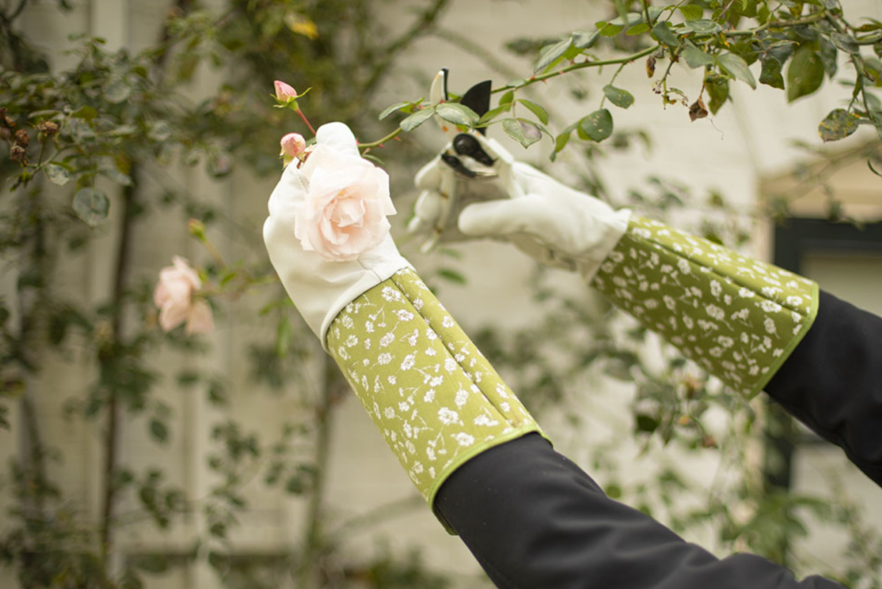 Ogrodnik w zielonych rękawiczkach w kwiaty wykorzystuje Małe grabki ogrodowe - stalowe i bukowe - do pielęgnacji jasnoróżowych róż w bujnym ogrodzie.