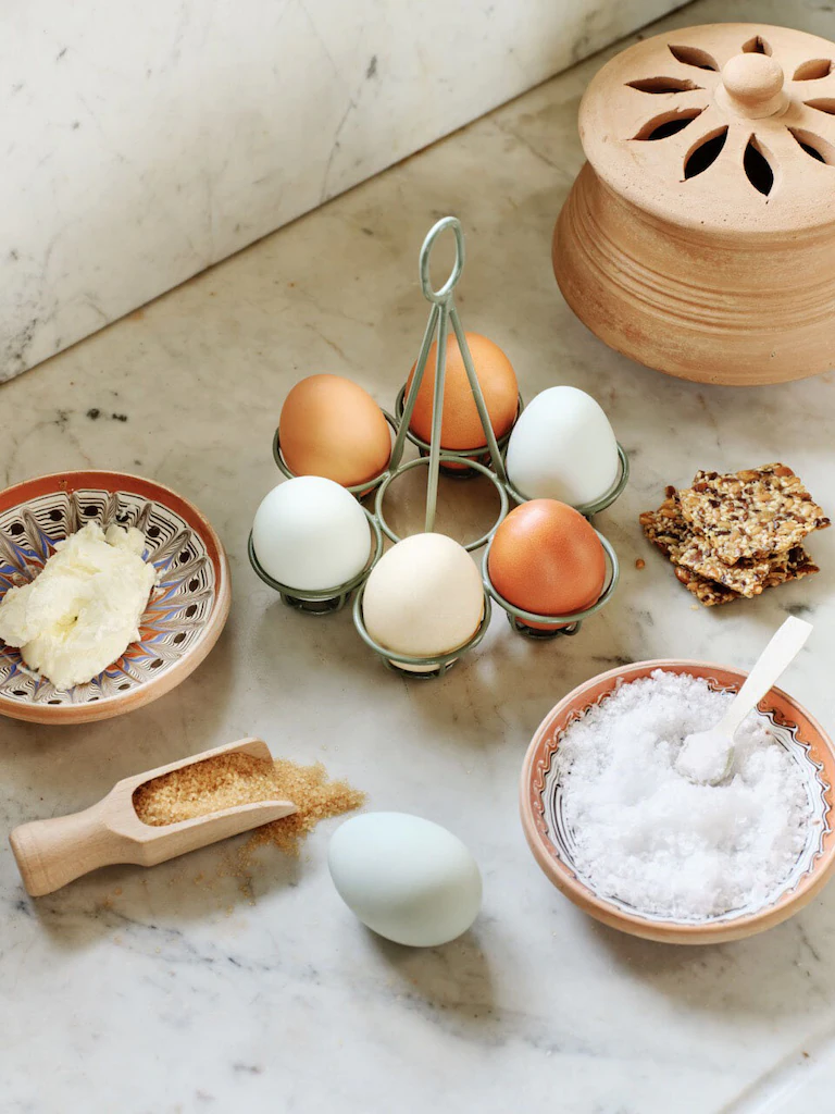 Jajka w zielonym metalowym stojaku na jajka, masło, brązowy cukier, sól, krakersy z nasionami i ceramiczny słoik na marmurze w butiku ogrodowym.