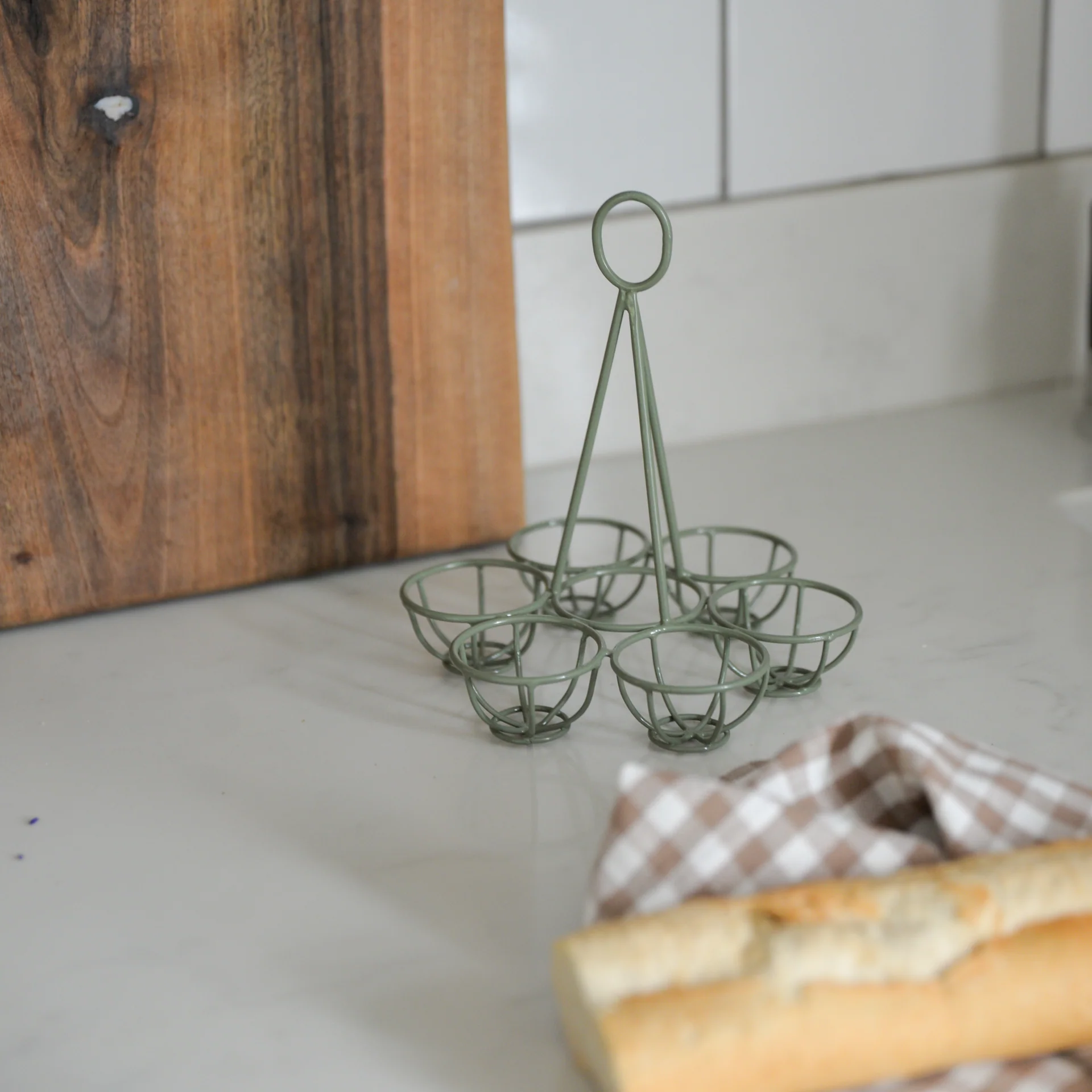 Stylowy stojak na jajka w szałwiowej zieleni na blacie, obok deski do krojenia; kawałek bagietki i kraciasta ściereczka.