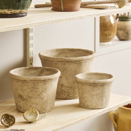 Zestaw trzech beżowych doniczek Osłonka z cementu Athena na drewnianej półce, w otoczeniu ceramicznych wazonów i narzędzi garncarskich w jasnym pomieszczeniu.
