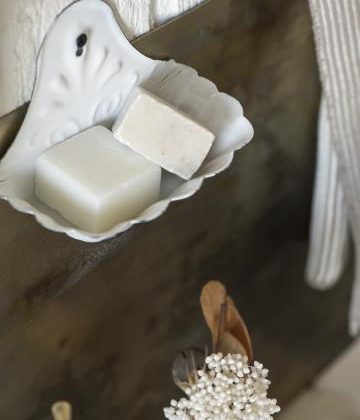 Biała, emaliowana, metalowa mydelniczka ścienna, zaprezentowana wraz z dwoma naturalnymi, kwadratowymi kostkami mydła — jedną w kolorze złamanej bieli, a drugą w kolorze beżowym — umieszczona nad suszonymi kwiatami i dekoracjami o tematyce ogrodowej; idealna do łazienek w stylu botanicznym lub ekspozycji w butikach ogrodniczych.